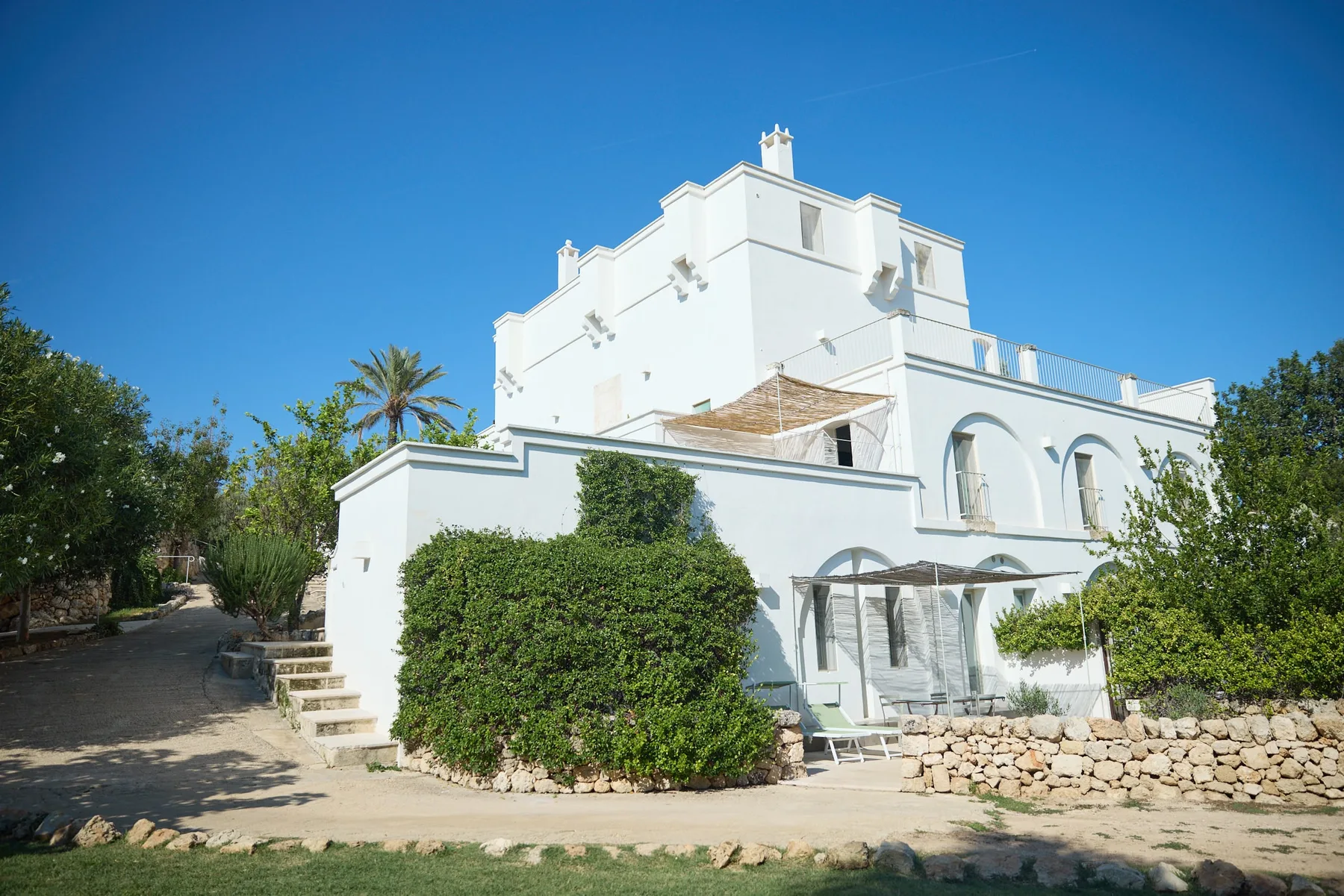 La facciata bianca di Masseria Alchimia tra la vegetazione, sotto il cielo blu