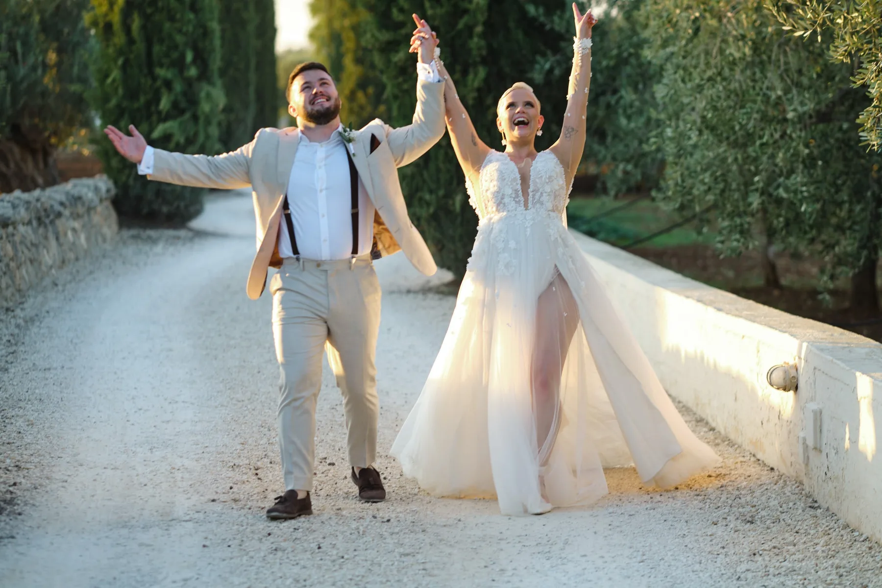 Natasha and Michael celebrating with their arms raised on the cypress-lined avenue at Masseria Alchimia