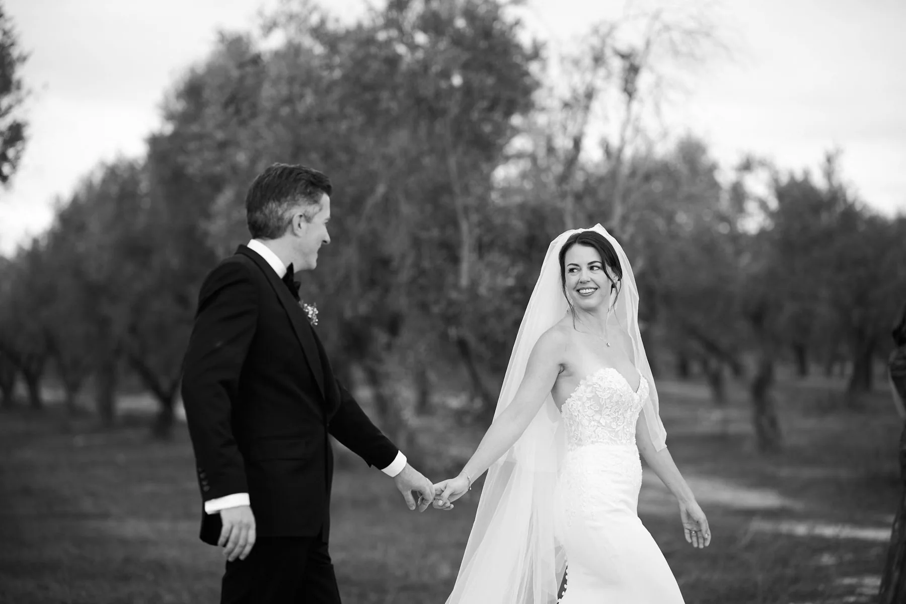 Katy and Gregory hand in hand among the olive trees, in black and white